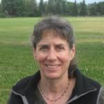 Woman with dark jacket and necklace smiling in front of green grassy field