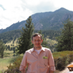 Man in salmon colored shirt standing in front of trees and mountains